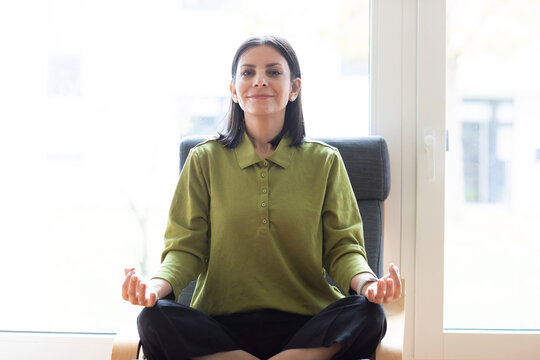 A woman sits cross-legged on a chair in front of a window, smiling and meditating indoors. Germany