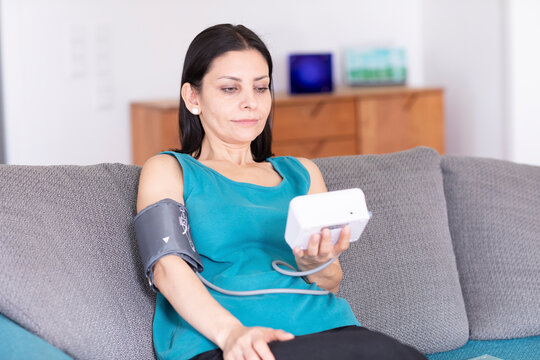 Woman sitting on a sofa measuring her blood pressure with a digital monitor. Germany