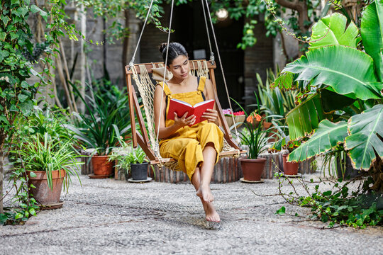Woman in yellow dress reading a book on a hanging swing in a lush green garden. Sierra Alhamilla, Almeria, Spain