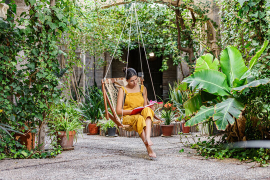 Woman reading on a swing in a lush green garden surrounded by plants and sunlight. Sierra Alhamilla, Almeria, Spain - Powered by Adobe