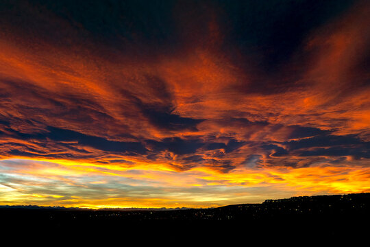 Vibrant fiery sunset with dramatic clouds over a silhouetted landscape and city lights. Calgary, Canada