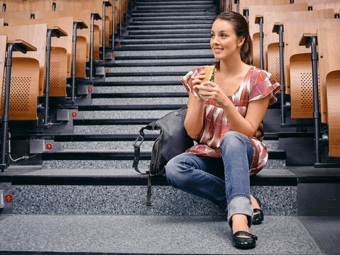 Young woman sitting on lecture hall steps, eating a sandwich, backpack beside her. Germany