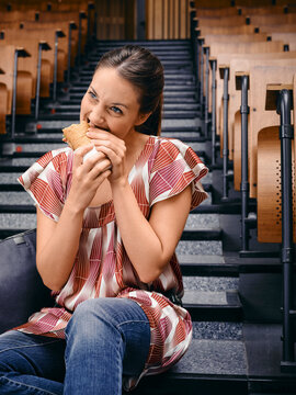 Young woman sitting on stairs in a lecture hall, enjoying a sandwich with a playful smile. Germany