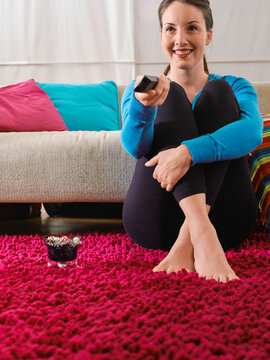 Woman sitting on a red rug in a living room holding a remote control with a smile. Germany