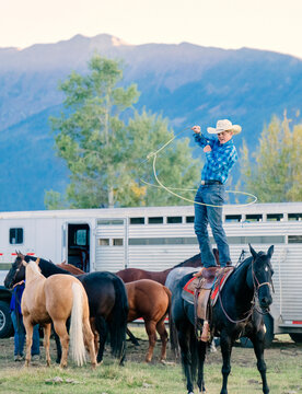 Cowboy standing on horseback twirling rope, surrounded by horses and trailer, mountain backdrop. Oregon, USA