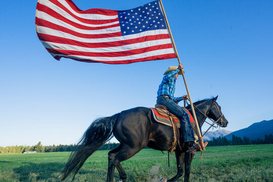 A cowboy rides a horse on a green field, holding a large American flag under a clear blue sky. Oregon, USA