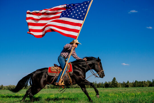 Cowboy on horseback rides swiftly, carrying a large American flag in a grassy field. Oregon, USA