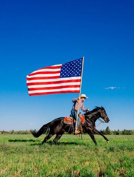 Cowboy riding a horse holding a large American flag in a grassy field under a clear blue sky. Oregon, USA