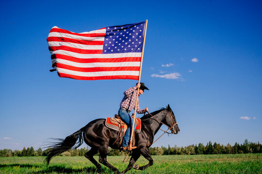 A person rides a black horse across a field waving a large American flag against a blue sky. Oregon, USA