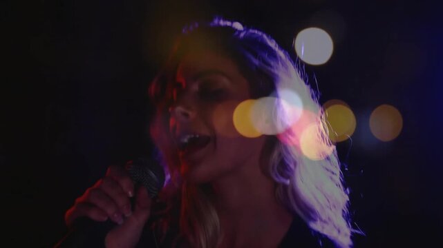 Female singer raising mic and singing, performing music on dim stage, bokeh lights crossing face