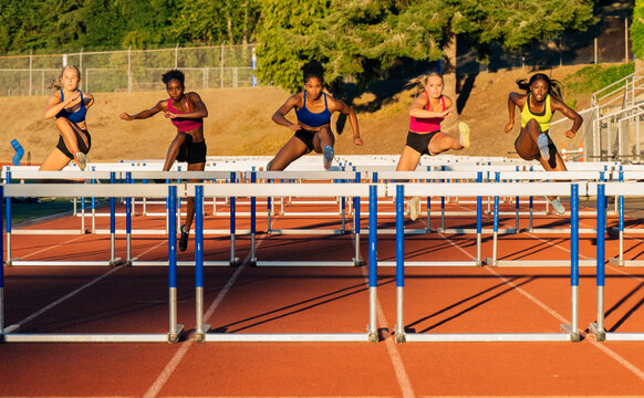 Five female athletes in colorful sportswear compete in a hurdle race on a sunny track. WA, USA