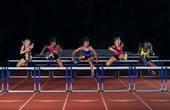Four athletes in vibrant sportswear leap over track hurdles in a synchronized motion. WA, USA