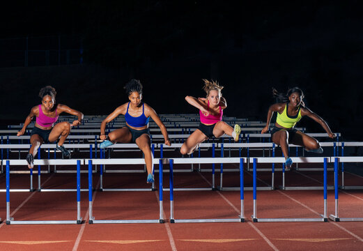 Four athletes in vibrant sportswear leap over track hurdles in a synchronized motion. WA, USA