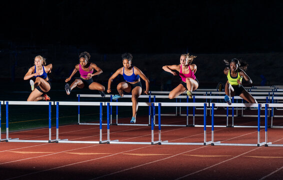 Five athletes in sportswear compete in a nighttime hurdle race on a track. WA, USA