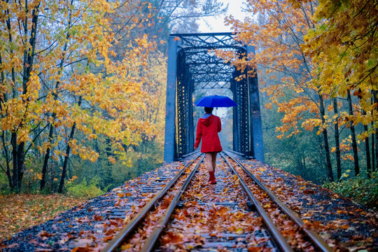 Woman with blue umbrella walking on railway tracks through a forest in autumn. WA, USA