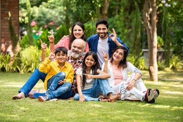 Happy Indian family of six sitting on lawn and pointing to present something outdoors.