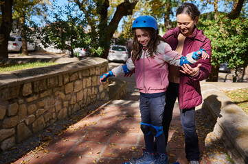 Mother Helps Daughter Balance On Park Path With Helmet And Protective Gear