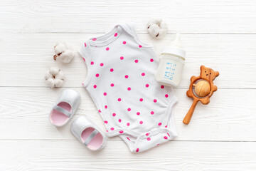Baby bodysuits and eco friendly accessories - booties, wooden rattle toy and baby bottle with milk on white wooden background, top view