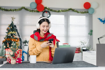 Happy Asian woman using smartphone with headphones in Christmas decorated office, holiday celebration at work