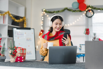 Young woman with headphones holding mug and smartphone at decorated office desk, cozy festive work celebration with laptop and gifts