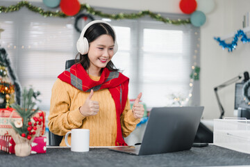 Young woman wearing headphones giving thumbs up during holiday video call, cozy sweater and festive...