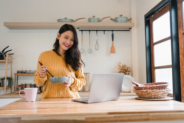 Young woman smiling while cooking in bright kitchen with laptop nearby, enjoying cozy morning and warm drink