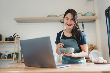 Young woman cooking in kitchen smiling while following online recipe, casual apron, cozy modern kitchen scene