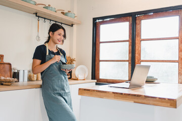Young woman smiling in apron eating cereal in bright kitchen while watching laptop