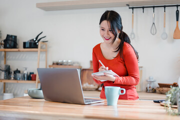 Young woman in red sweater smiling while taking notes and working on laptop in kitchen