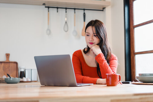 Young woman in red top working on laptop at kitchen table with coffee mug, thoughtful expression and natural light - Powered by Adobe