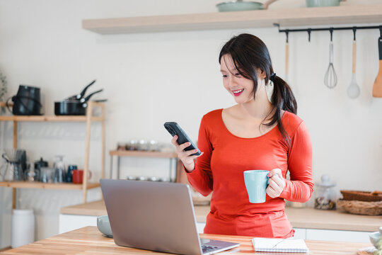 Young woman holding coffee mug and smartphone while working on laptop in kitchen, smiling and relaxed - Powered by Adobe