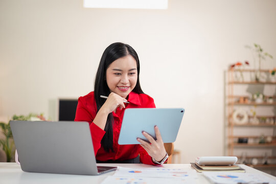 Young woman in red blouse working on tablet and laptop at home office smiling with stylus and paperwork