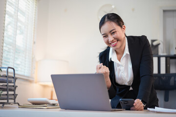 Young businesswoman celebrating success while working on laptop and holding smartphone in bright office