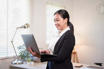 Young woman holding laptop smiling while working in bright home office with plants and lamp