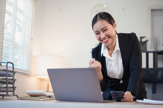 Young businesswoman celebrating success while working on laptop and holding smartphone in bright office - Powered by Adobe