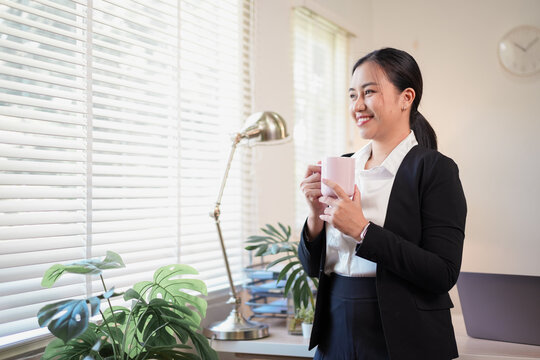 Woman in business suit holding mug and smiling by bright office window, relaxed morning mood