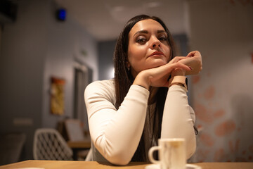 Young woman with long dark hair sits at a table, hands clasped, in a cozy cafe setting, warm lighting creating an inviting atmosphere for conversation and relaxation