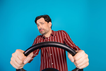 Young handsome guy driving steering wheel posing with puzzled expression against blue background wearing a casual striped shirt and stylish fashion vibe