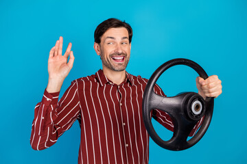 Young man with steering wheel smiling and waving hand against blue background promoting fun driving lifestyle and stylish casual fashion