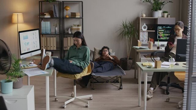 Long shot of three relaxed Asian coworkers using digital devices while having break in modern office workspace