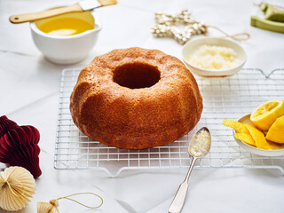 A freshly baked lemon bundt cake rests on a metal rack. Glaze ingredients, lemon zest, and melted butter are nearby, preparing for decorating. Christmas ornaments create a festive atmosphere