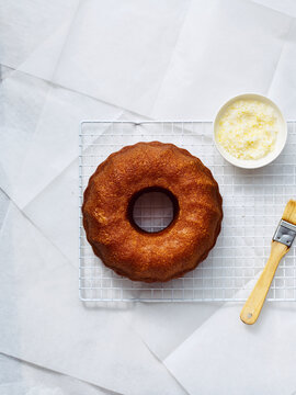 A golden brown bundt cake cools on a white wire rack. Next to it is a bowl filled with a light colored glaze and a wooden pastry brush, all on white parchment