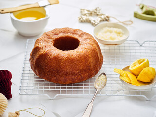 A freshly baked lemon cake cools on a rack. Butter, lemon zest, lemon slices, and holiday decorations are on a white table, suggesting a festive baking scene