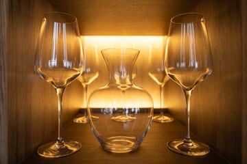 Set of wine glasses and a glass decanter arranged on a wooden shelf under warm backlighting in a...