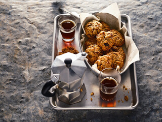 A metal tray displays homemade oatmeal raisin cookies in parchment paper, two glasses of dark coffee, and a classic stovetop Moka coffee maker. Ready for morning enjoyment