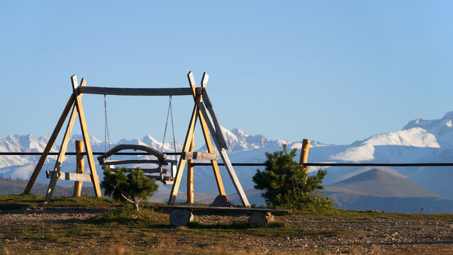A wooden swing sits on a steep mountainside against a backdrop of rocky, snow-capped mountains.