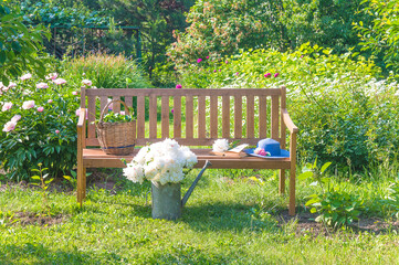 Wicker basket with plants, open book and blue hat on a wooden bench in blooming summer garden by sunny day