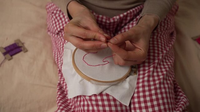 Overhead view of feminine hands embroidering two connected hearts with vibrant red thread on white fabric held in a wooden hoop, cozy handmade needlework craft project