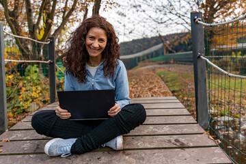 Woman smiling while using a laptop outdoors on a wooden bridge during autumn