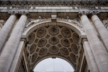 Low-angle view of the ornate ceiling and columns of the Cinquantenaire Arch in Brussels, Belgium. The image highlights the intricate geometric patterns, classical sculptures, and crafmanship